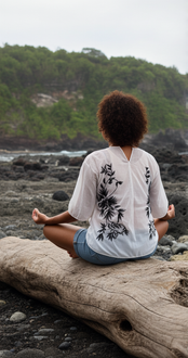 A person sits in a meditative pose on a large piece of driftwood on a rocky beach, facing the ocean.