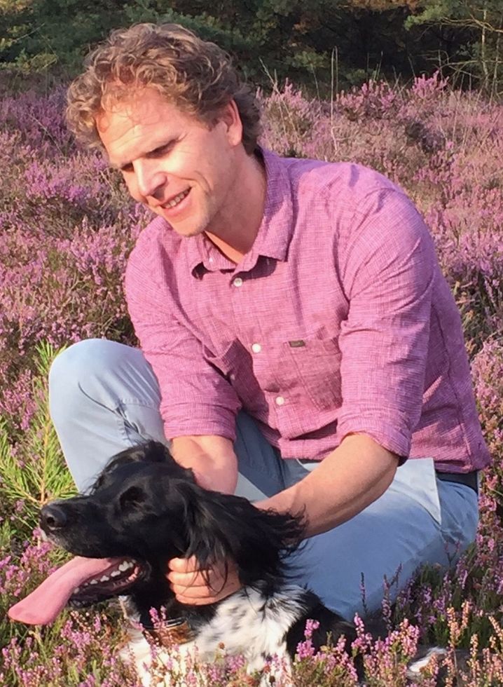Man in roze shirt en blauwe spijkerbroek aait een zwart-witte hond in een veld met paarse bloemen.