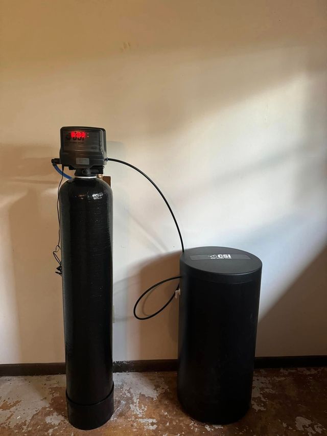 Black water softener and salt tank connected by a hose, standing against a beige wall on a hardwood floor.