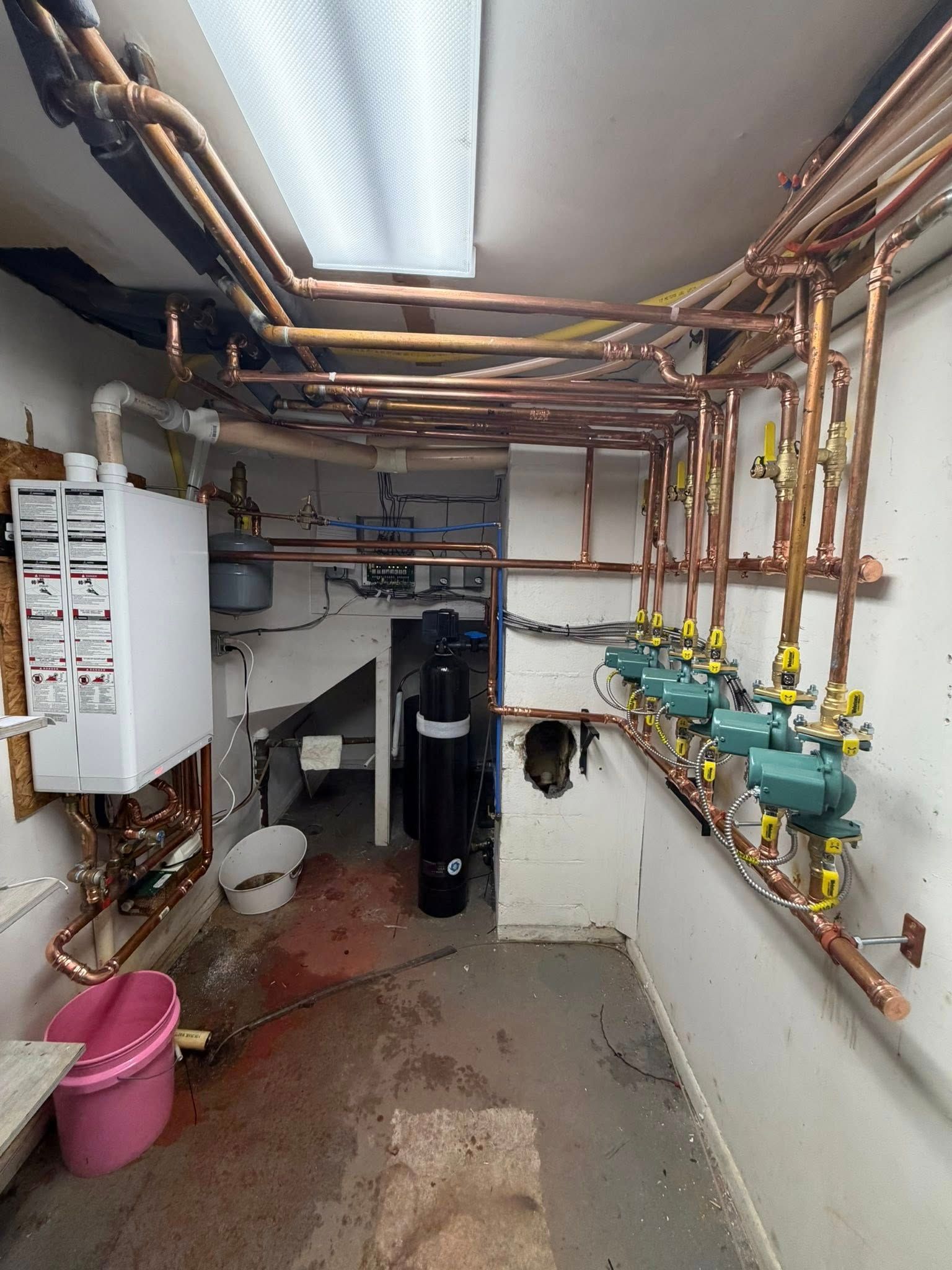 Utility room with exposed copper pipes, water meters, and cleaning buckets on a concrete floor.