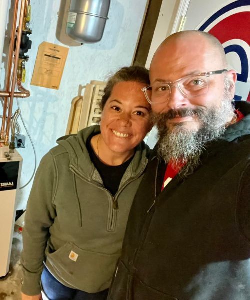 Two people smiling indoors, wearing casual jackets, standing close together near pipes and equipment.