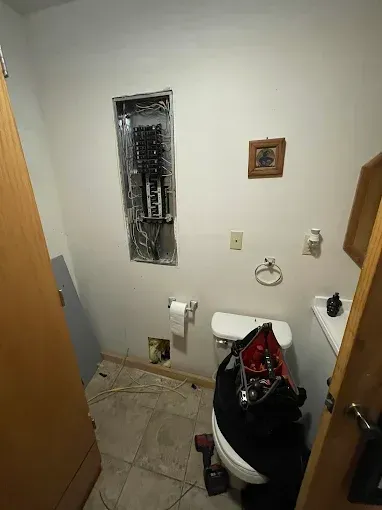 Small bathroom with exposed electrical panel, toilet, sink, and scattered tools or supplies during ремонт.