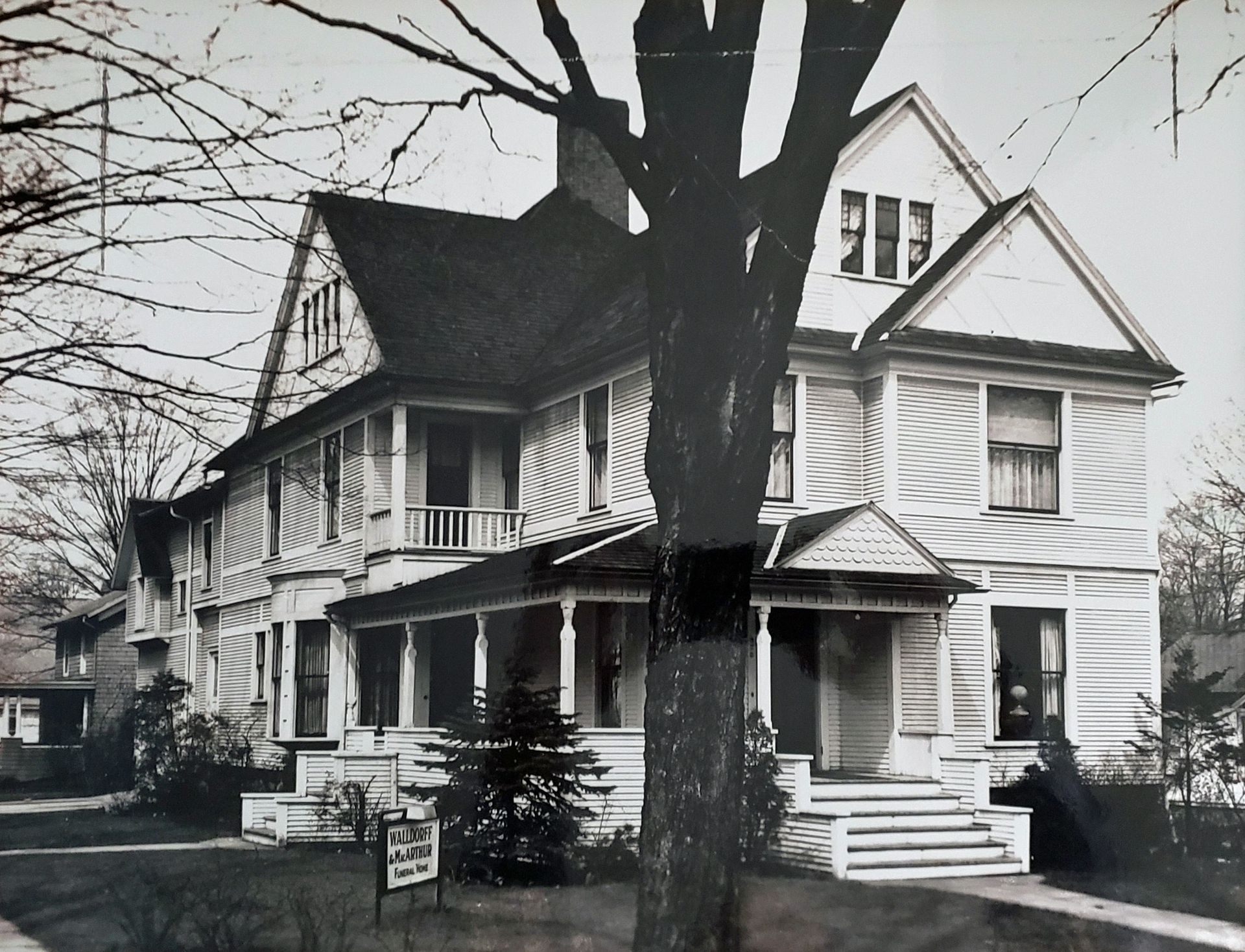 Vintage black and white photo of a two-story house with a porch and tree in front.