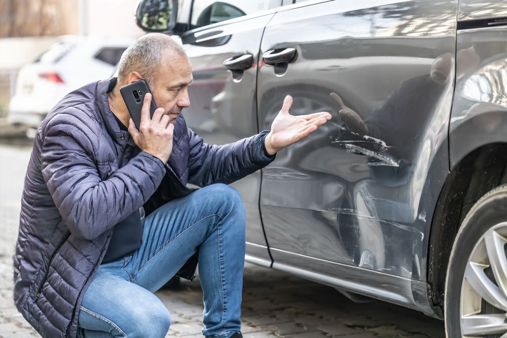 Een man knielt naast een beschadigde auto terwijl hij belt op een mobiele telefoon.