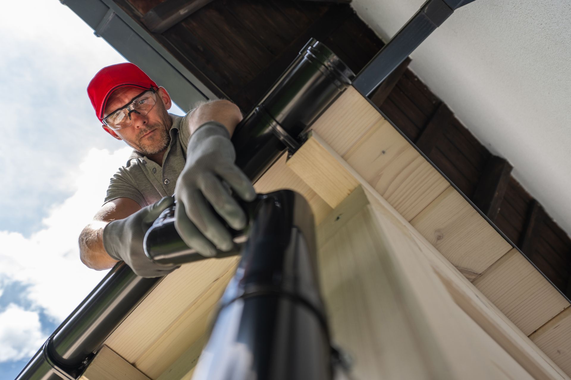 A person in a red cap, safety glasses, and gloves installs a black gutter section on the corner of a building exterior.