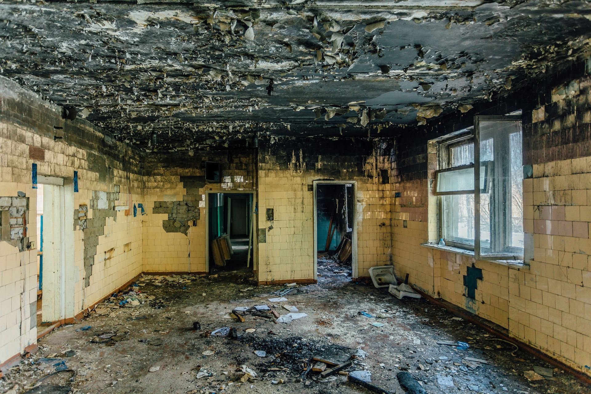 An abandoned, dilapidated room with fire-damaged, black ceilings, peeling walls, and debris scattered across the floor.