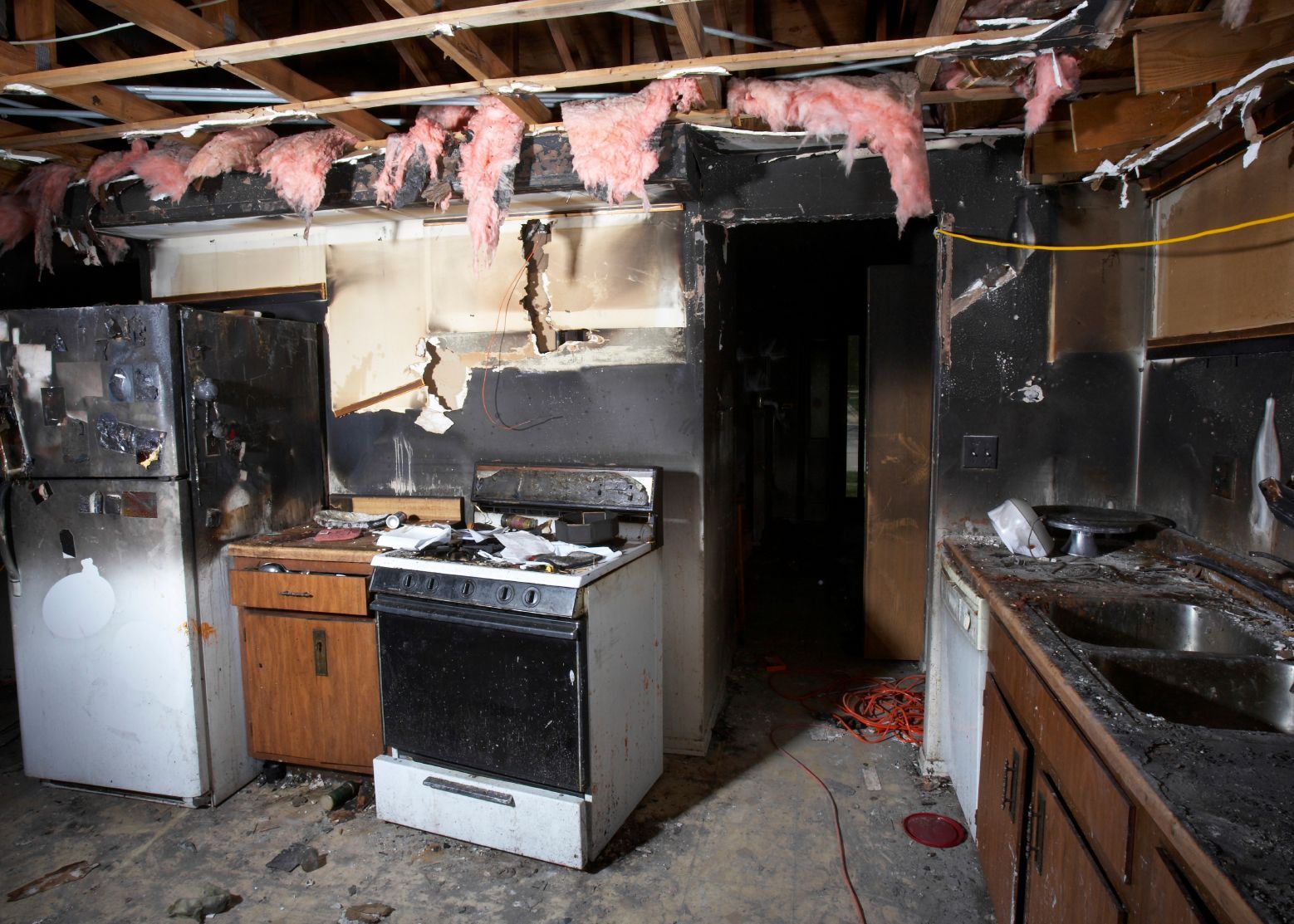 A kitchen after a fire, showing smoke-damaged walls, an exposed ceiling with insulation, and charred appliances.