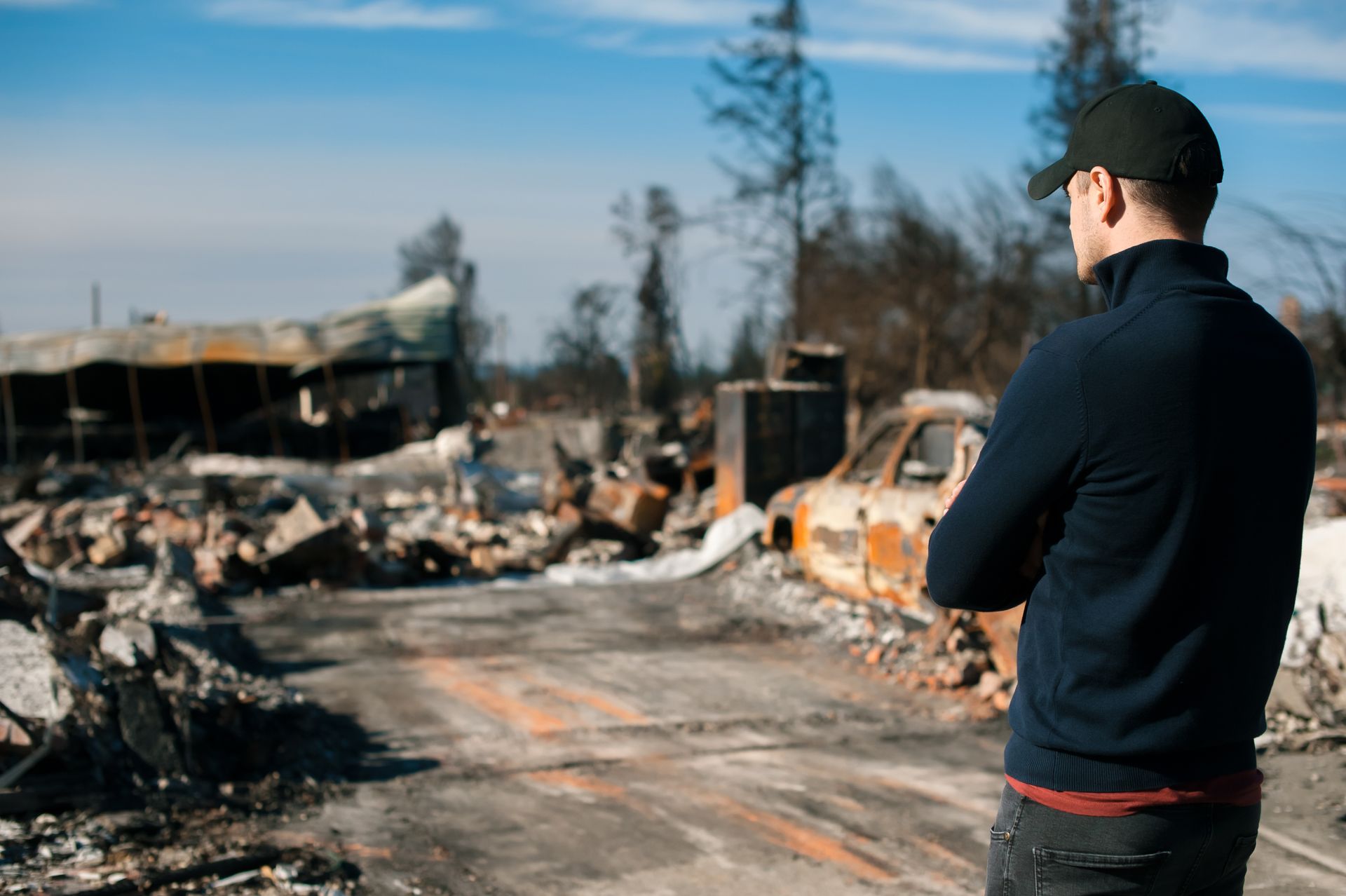 A person in a dark sweater and cap stands with arms crossed, looking at the charred ruins of a building and a burnt car.