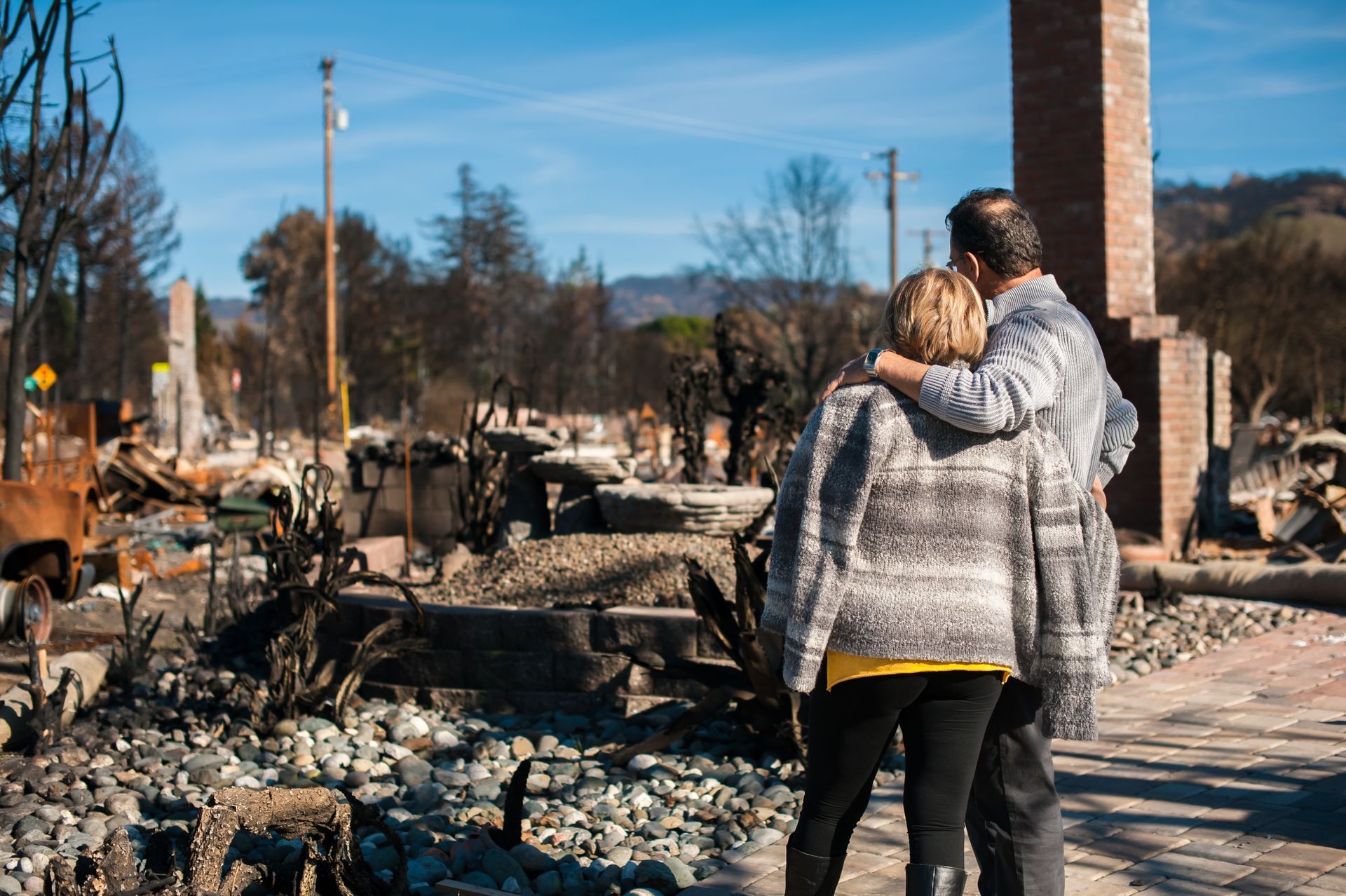 A couple stands with arms around each other, looking at the charred ruins of a building destroyed by wildfire.