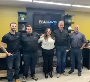 Équipe de cinq personnes Paul Davis debout dans un bureau, souriant devant un écran avec logo.