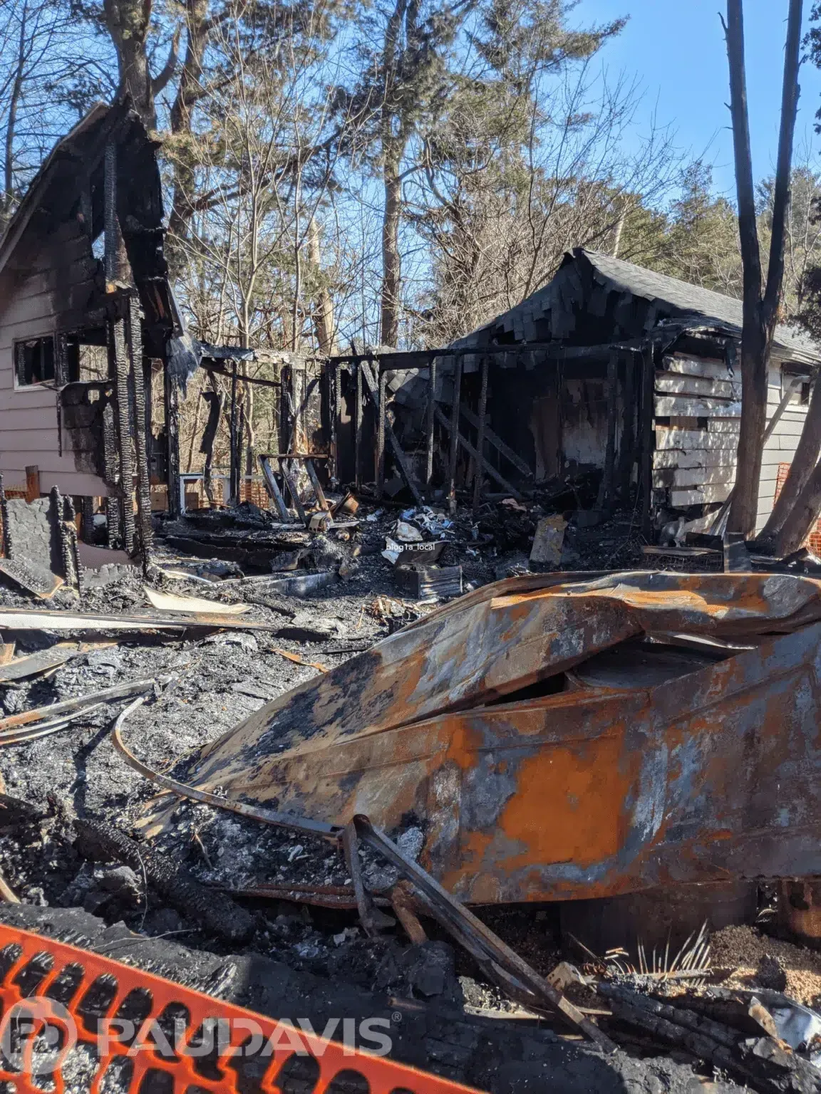 Severely fire-damaged home exterior with collapsed walls and burned debris in wooded area.