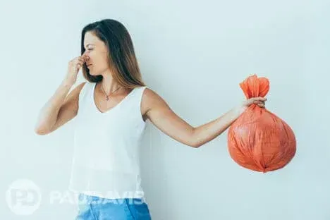 Woman holding a smelly red garbage bag at arm’s length while pinching her nose in discomfort.