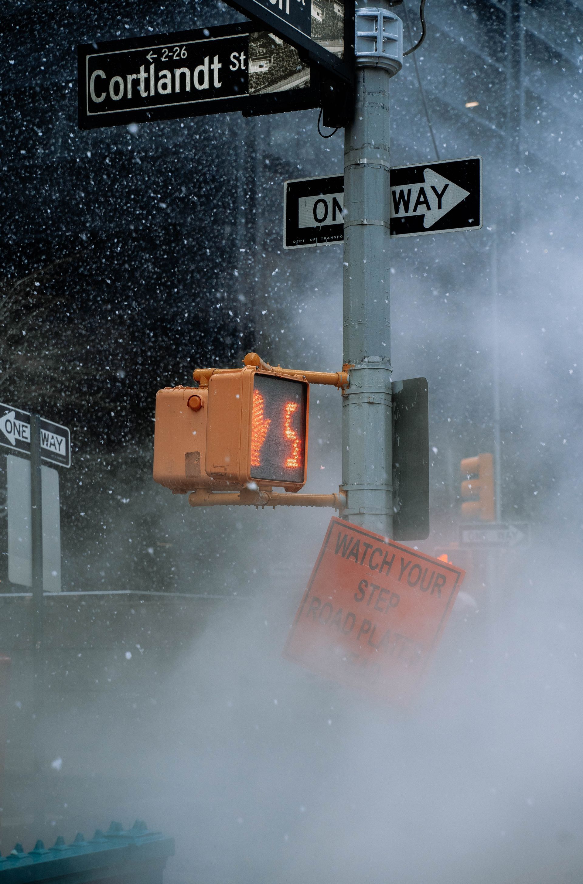 Snowstorm at city crosswalk with stop signal, street signs, and steam reducing visibility.
