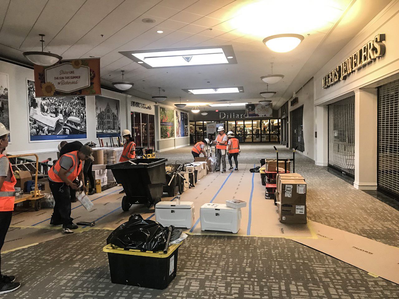 Construction workers in safety vests work in a shopping mall hallway with cardboard floor protection and equipment.