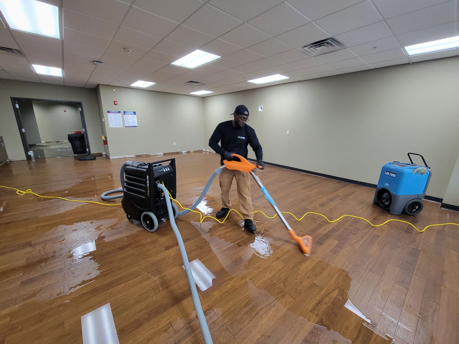 A person in work clothes uses professional extraction equipment to remove standing water from a laminate floor in an office.