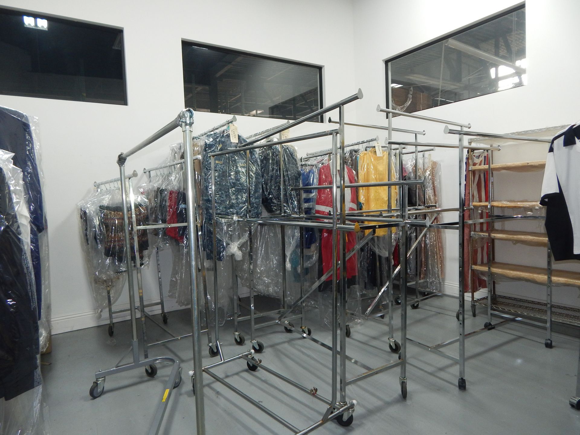 Rolling garment racks filled with clothing, some in plastic covers, in a clean, minimalist storage room with white walls.