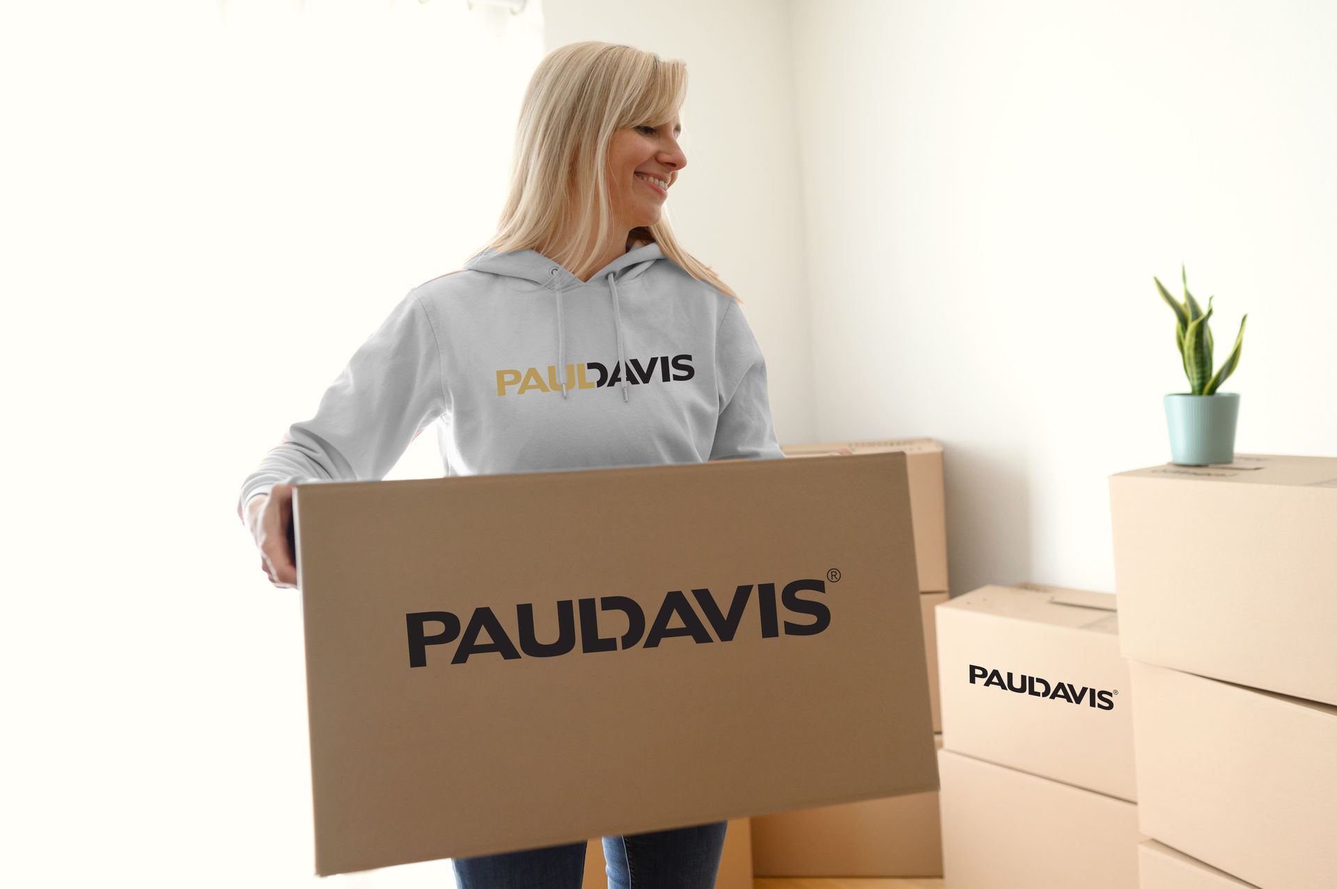 A smiling person in a gray Paul Davis hoodie carries a cardboard box with the Paul Davis logo in a brightly lit room.