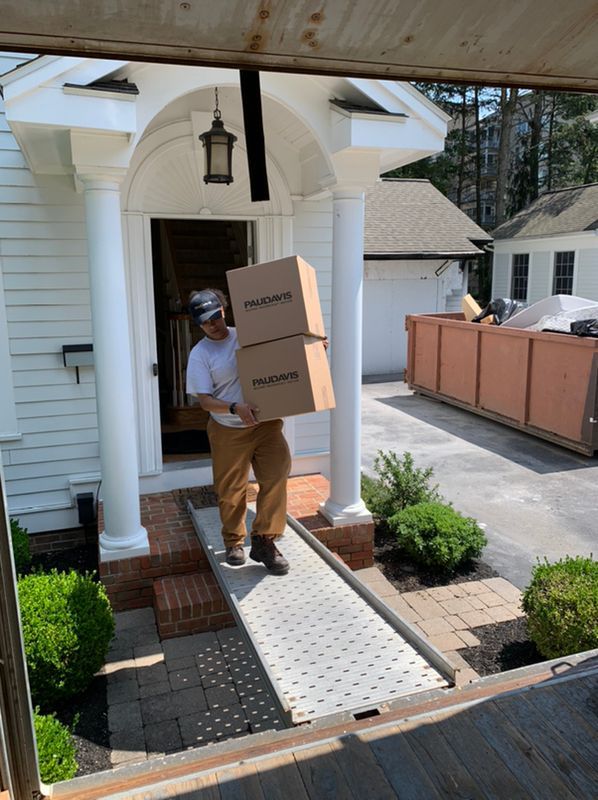A person carries two stacked boxes down a ramp from a white home entrance with columns and a nearby dumpster.