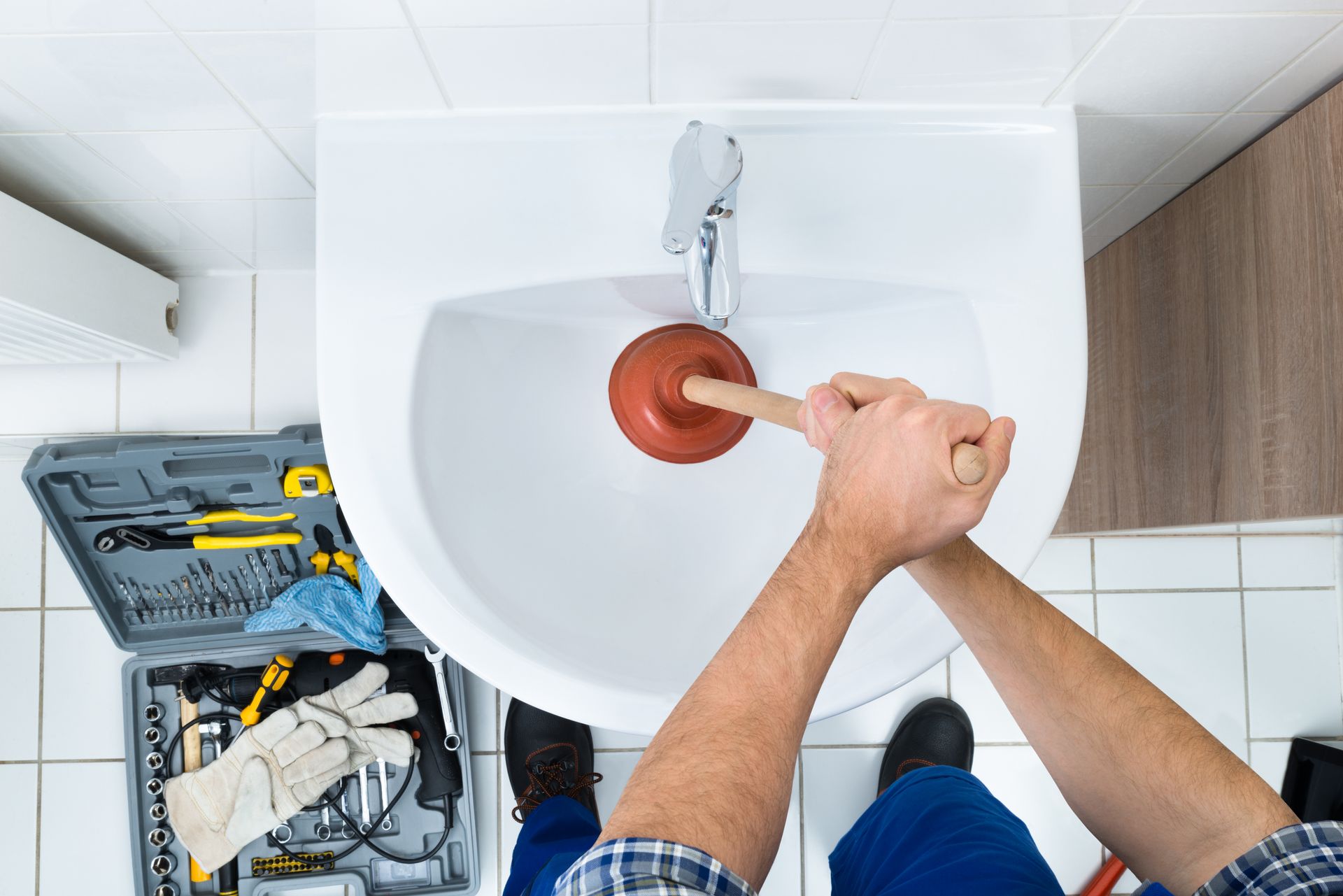 A person uses a red plunger to unclog a white bathroom sink, with a toolbox containing various tools nearby on the floor.
