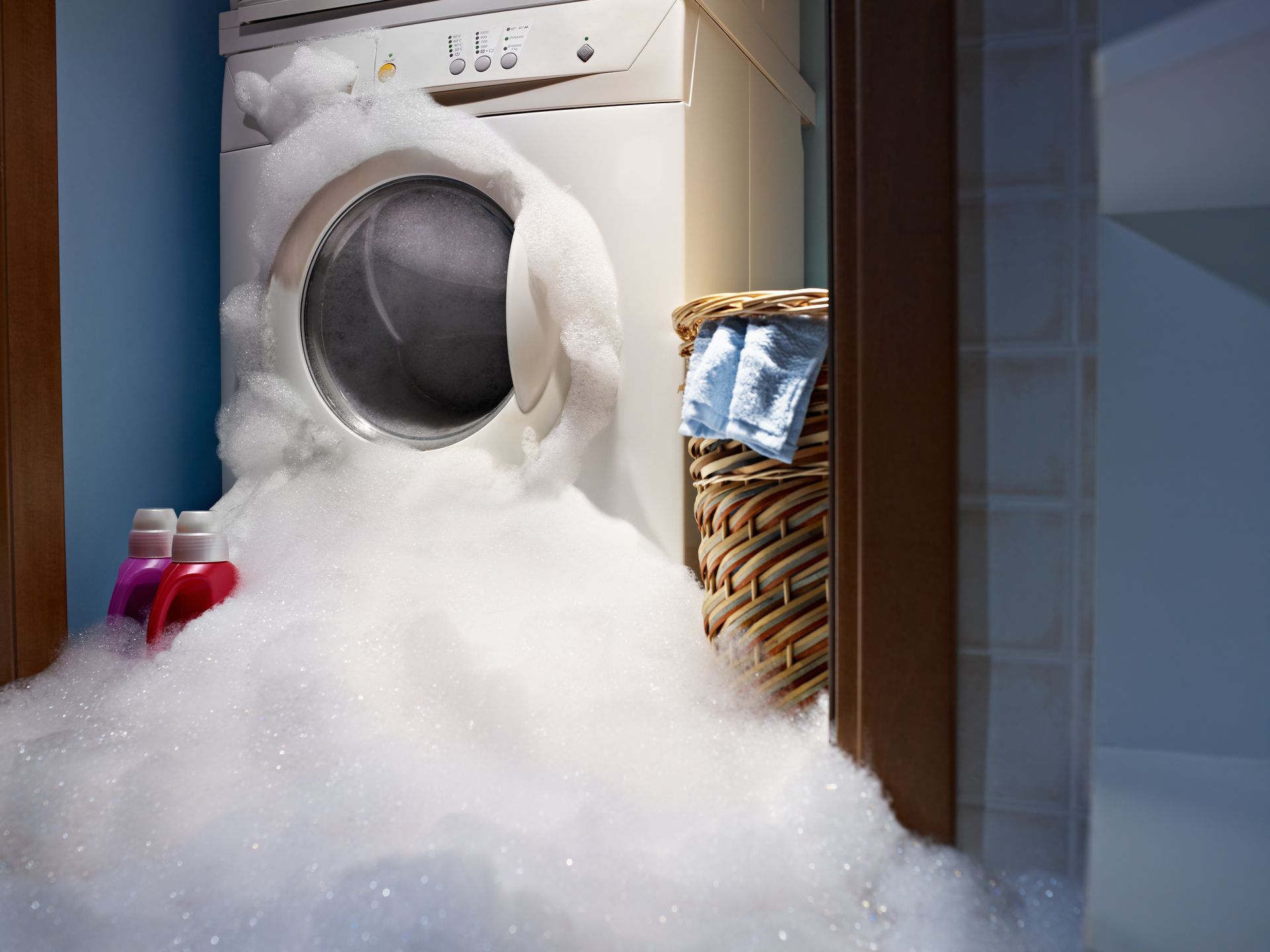 A washing machine overflowing with soapy suds into a laundry room, with detergent bottles nearby.