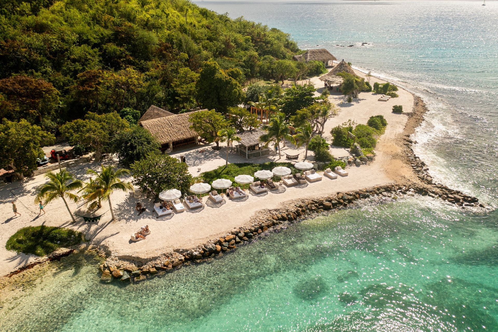 Aerial view of a white sand beach with turquoise water. Beach chairs, umbrellas, and lush trees line the shore.