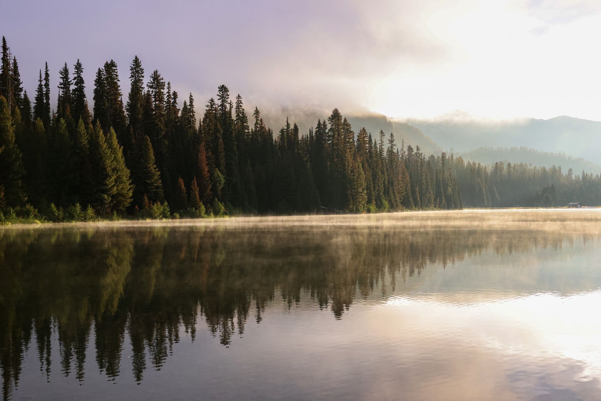 Still lake reflecting a forested shoreline and a hazy sky; sunlight.