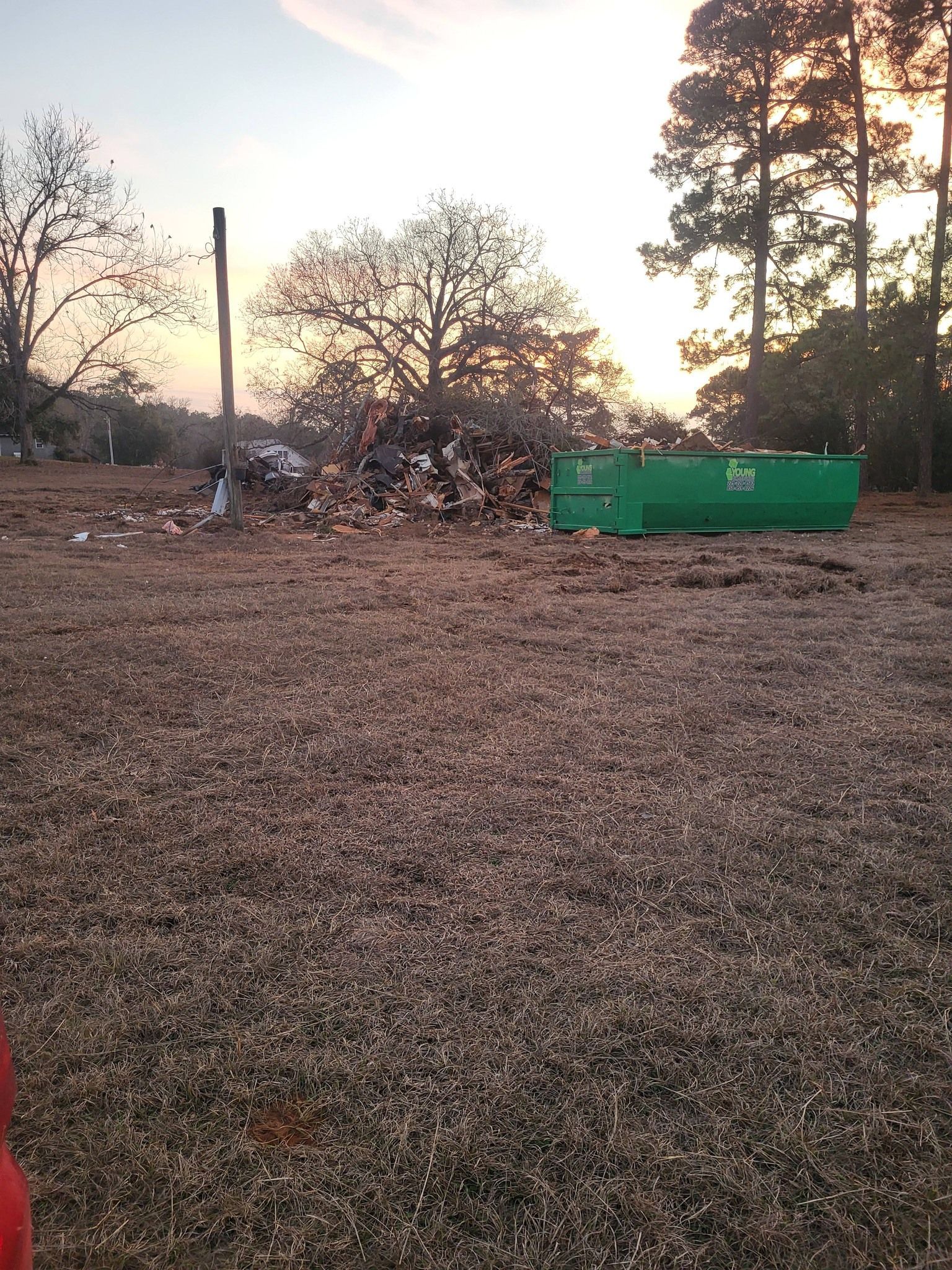 Pile of debris and green dumpster in a field at sunset, with trees in the background.