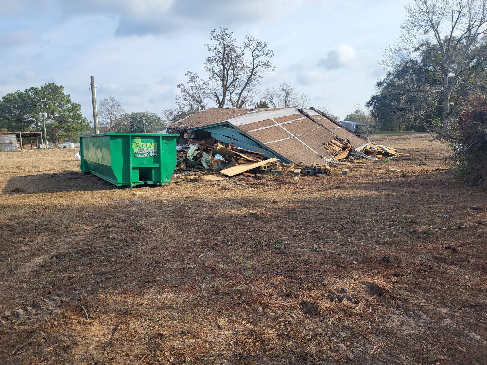 Demolished house with debris pile and a green dumpster in a field. Blue sky, brown grass.
