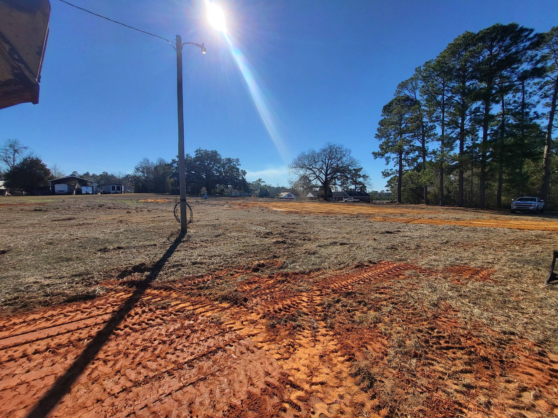 Open field with red dirt, utility pole, trees, and bright sun.