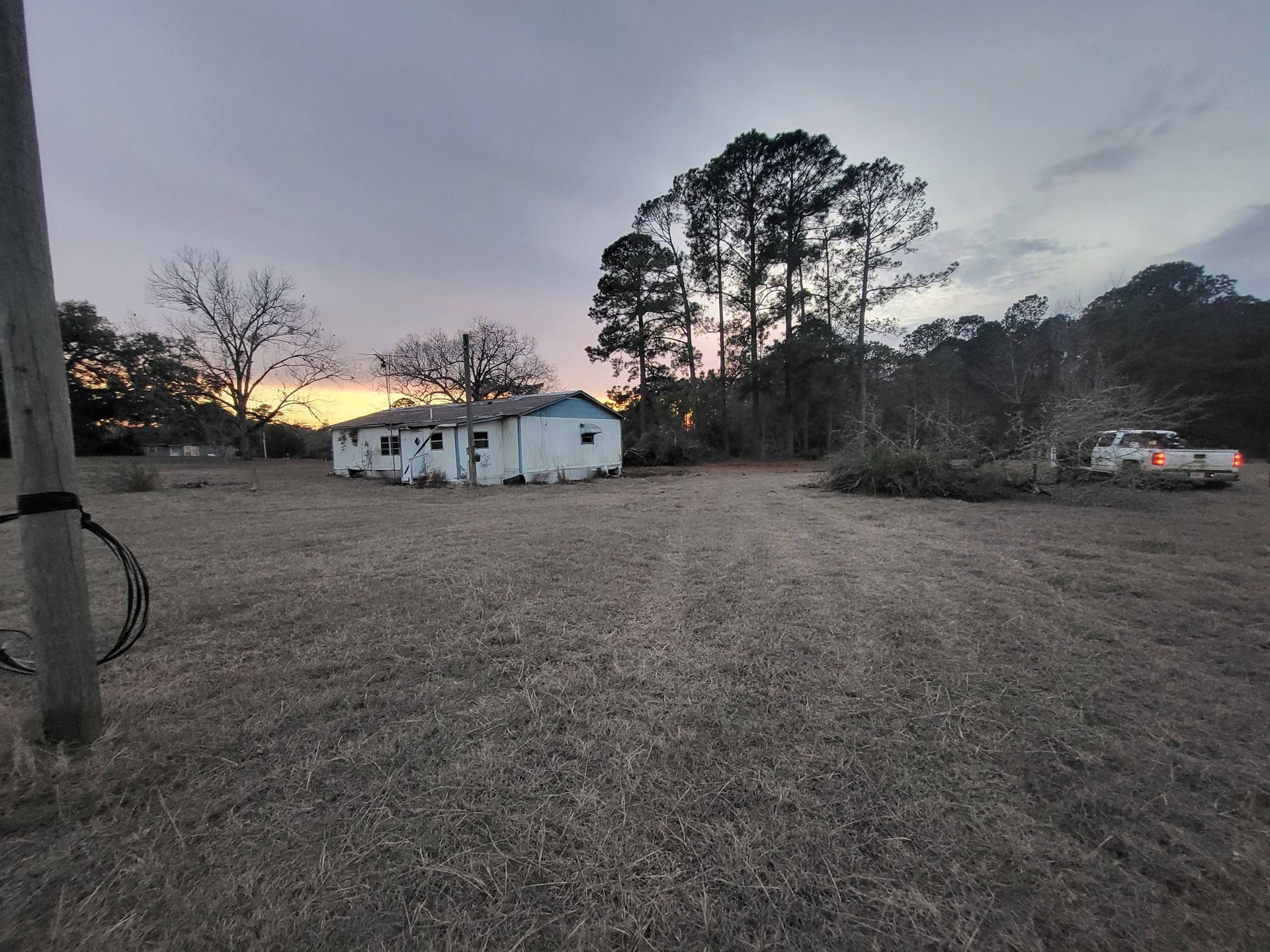 A small, weathered house at dusk, with tall trees and a utility pole in the foreground.