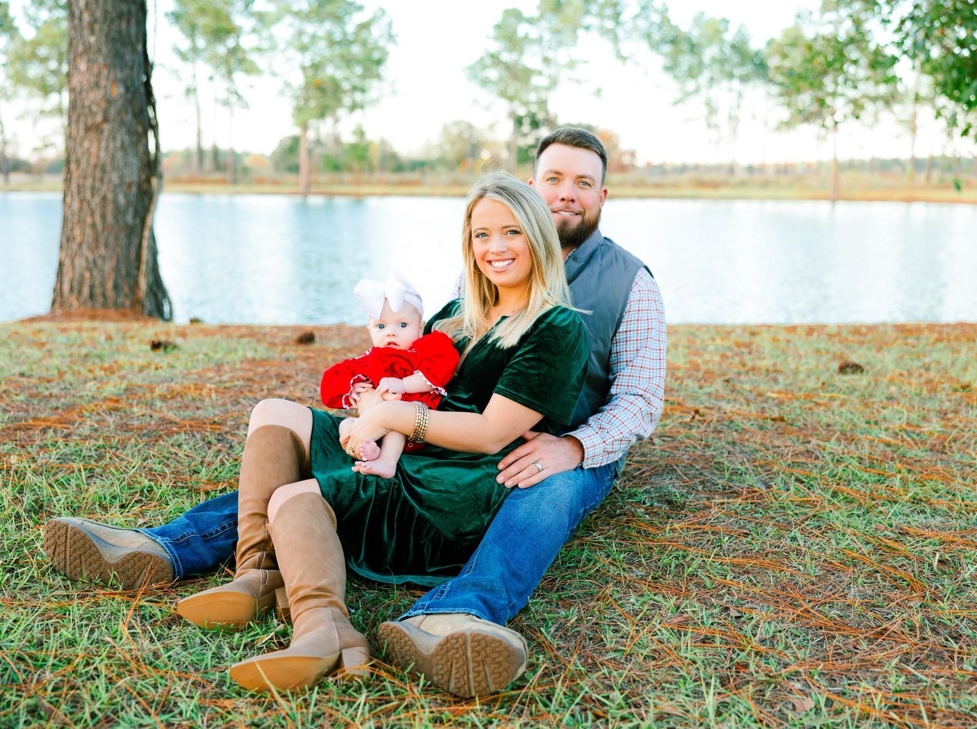 Family of three sitting on grass near lake; woman in green dress holding baby, man with beard.