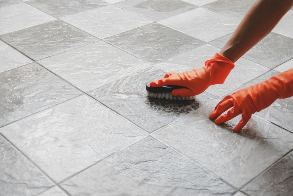 A Person Wearing Orange Gloves Is Cleaning A Tile Floor With A Brush — Lindfield’s Big Red Carpet Cleaning & Pest Control in Tuncurry, NSW