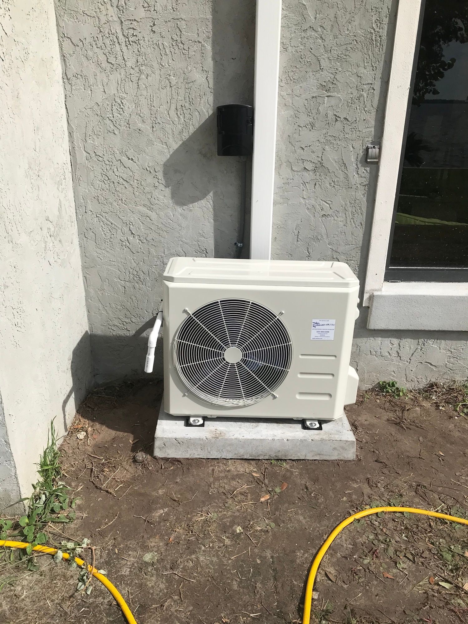 A white air conditioner is sitting outside of a building next to a yellow hose.