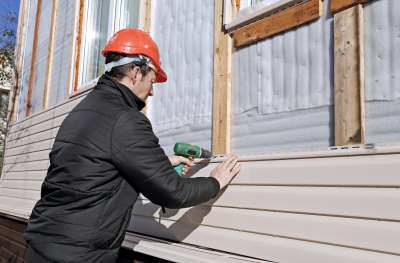 A man in a red safety helmet uses an electric screw driver to attach exterior cladding to a building
