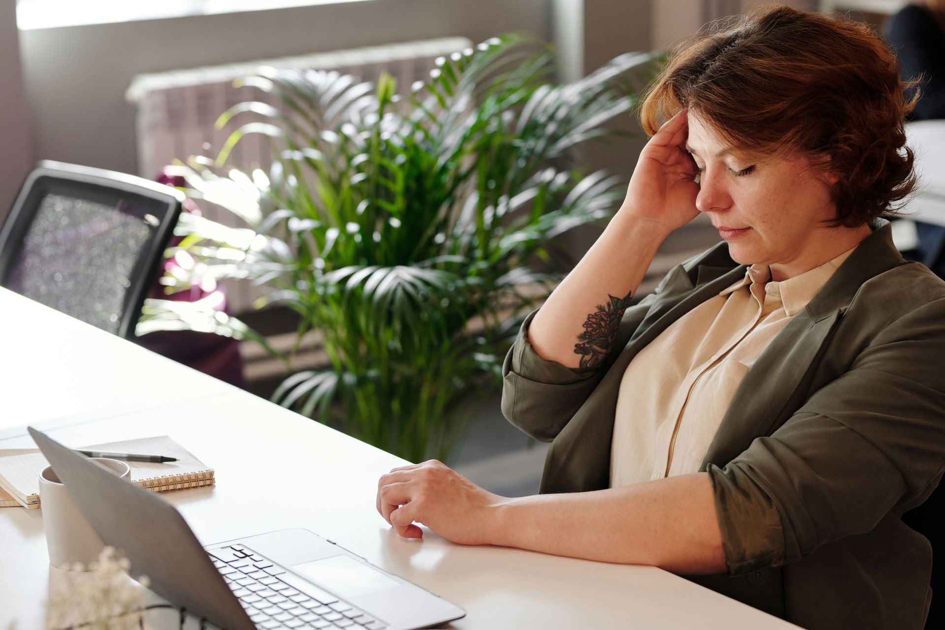 Person sitting at a desk, hand on forehead, working beside an open laptop in a bright office.
