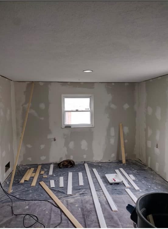 Room under renovation: drywall, window, wood trim on floor, tools. Gray walls and floor, white ceiling.