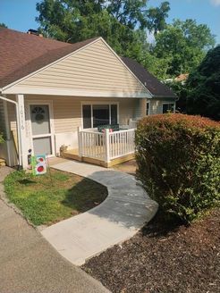 A small, beige house with a curved concrete walkway leading to a porch with white railings. A large green bush sits to the right.