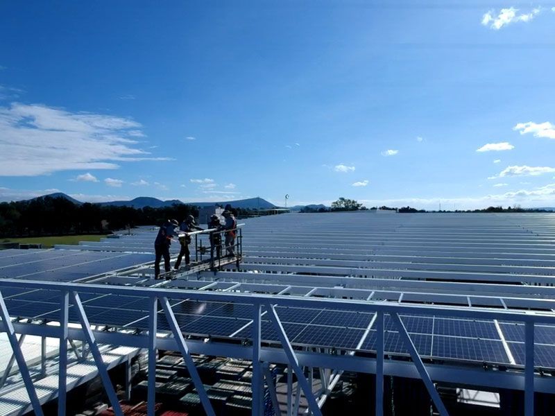 Obreros en una plataforma inspeccionando paneles solares en un tejado, con cielo azul y montañas al fondo.