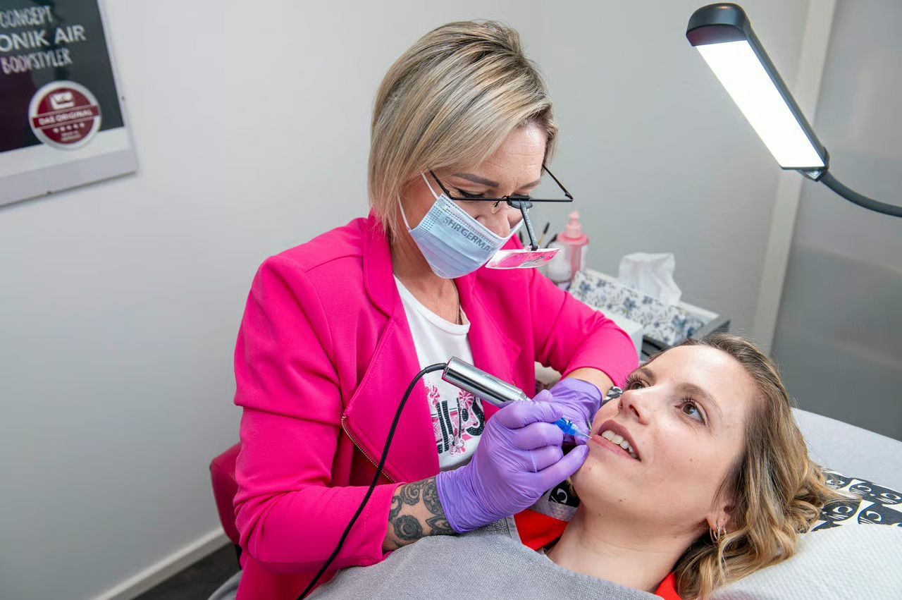 Eine Frau in einer rosa Jacke führt in einem Salon eine Permanent Make-Up bei den Lippen einer Kundin durch.