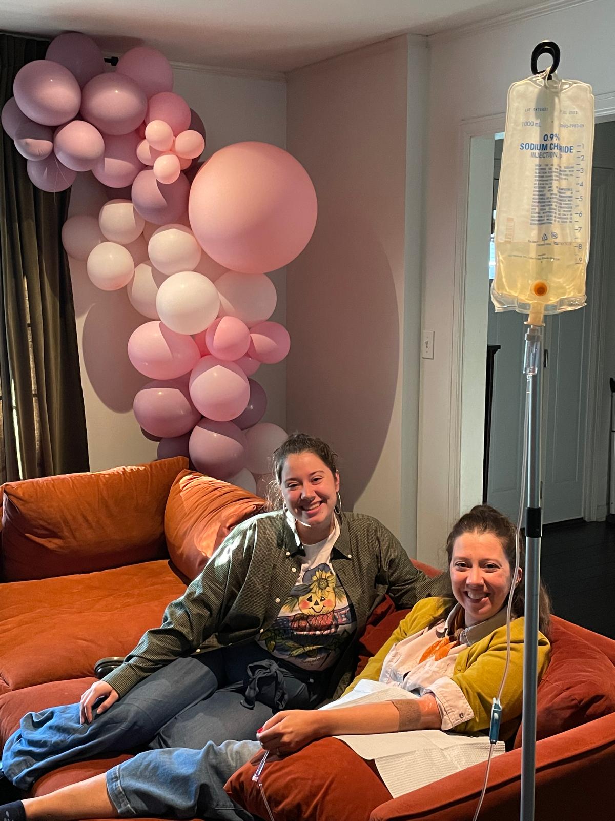 Two women are sitting on a couch with balloons in the background.