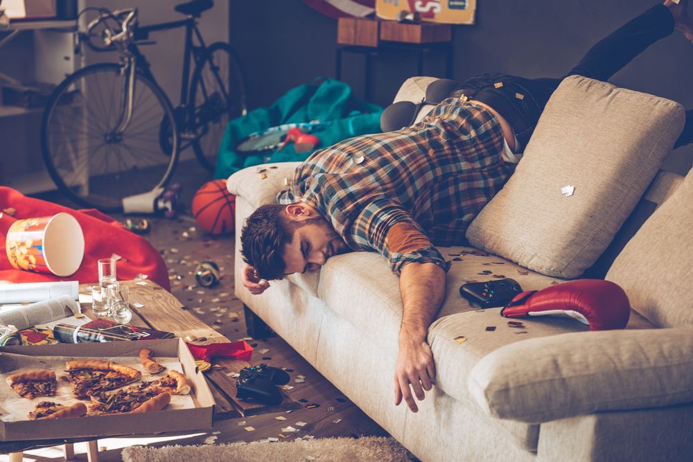 A man is sleeping on a couch in a messy living room.