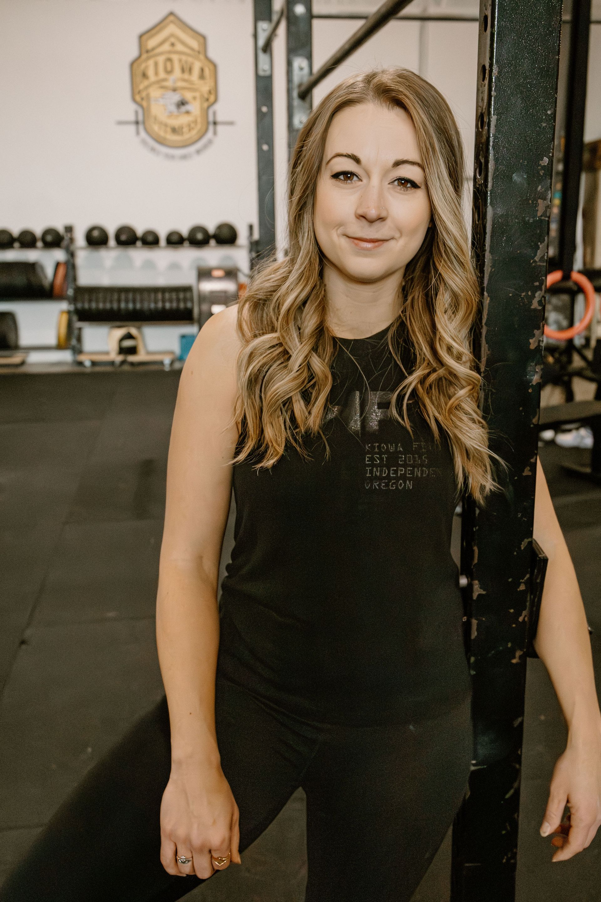 Woman in workout clothes leans against a weight rack in a gym, smiling.