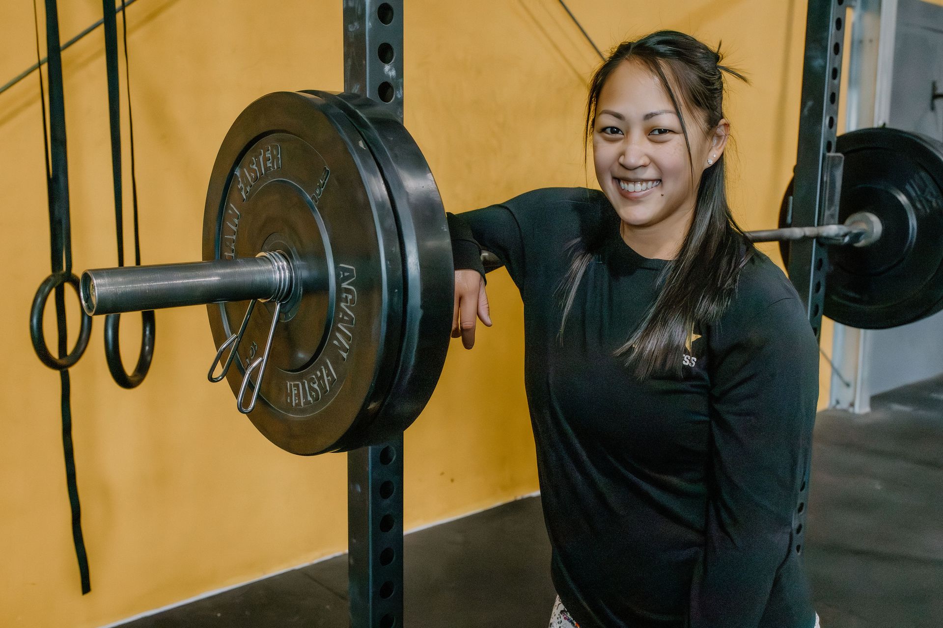 Woman in black shirt smiles, leaning on weight bar with plates in a gym. Yellow wall in background.