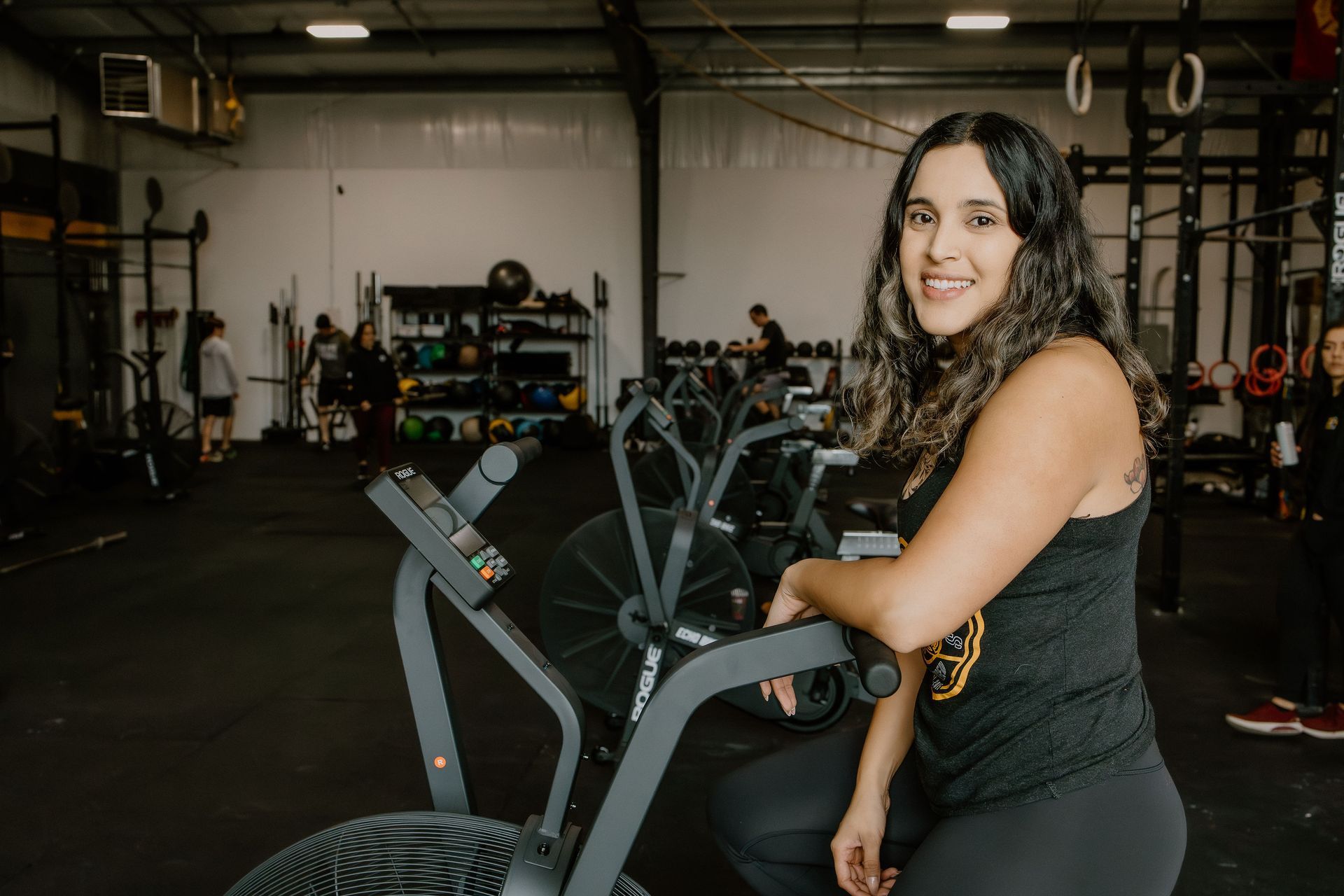 Woman smiling, leaning on an exercise bike in a gym, with other equipment and people in the background.