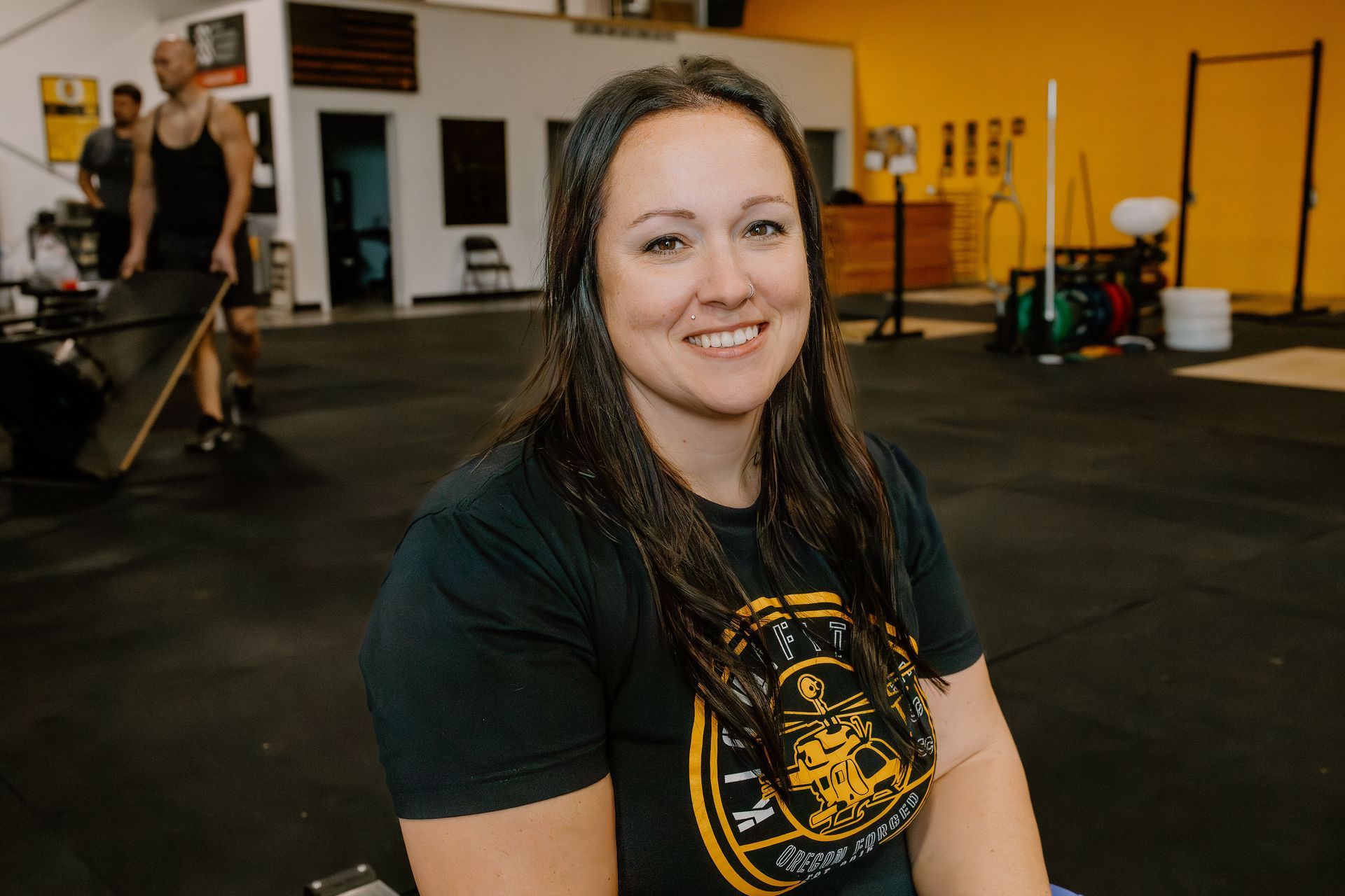 Woman smiles in a gym, wearing a black shirt with a gold graphic. Workout equipment visible in background.