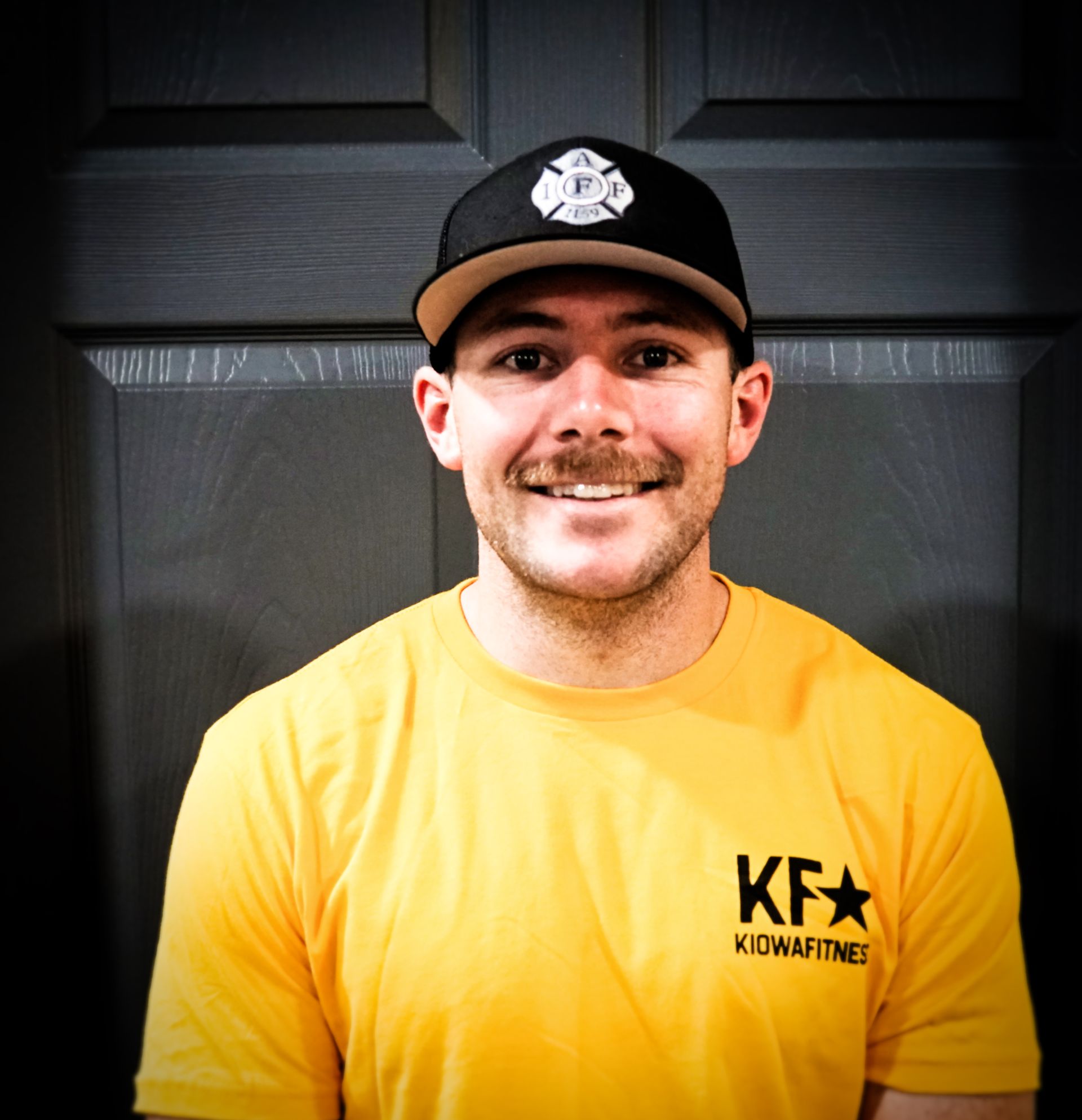 Man wearing a black hat and yellow shirt smiles at the camera, in front of a gray door.