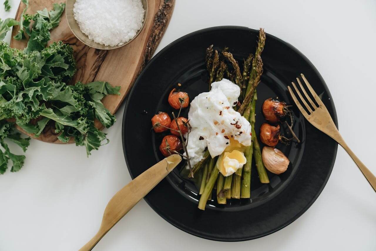Black plate with asparagus, tomatoes, poached egg, and gold utensils.