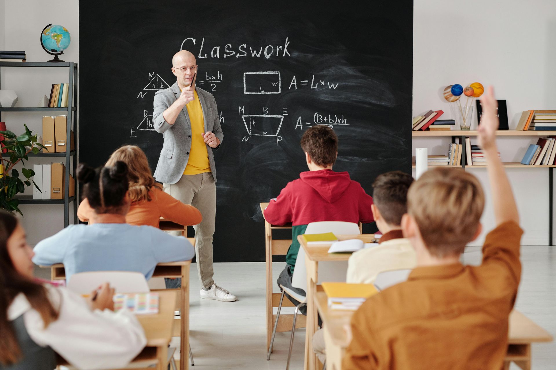 Teacher lecturing at chalkboard in classroom as student raises hand.