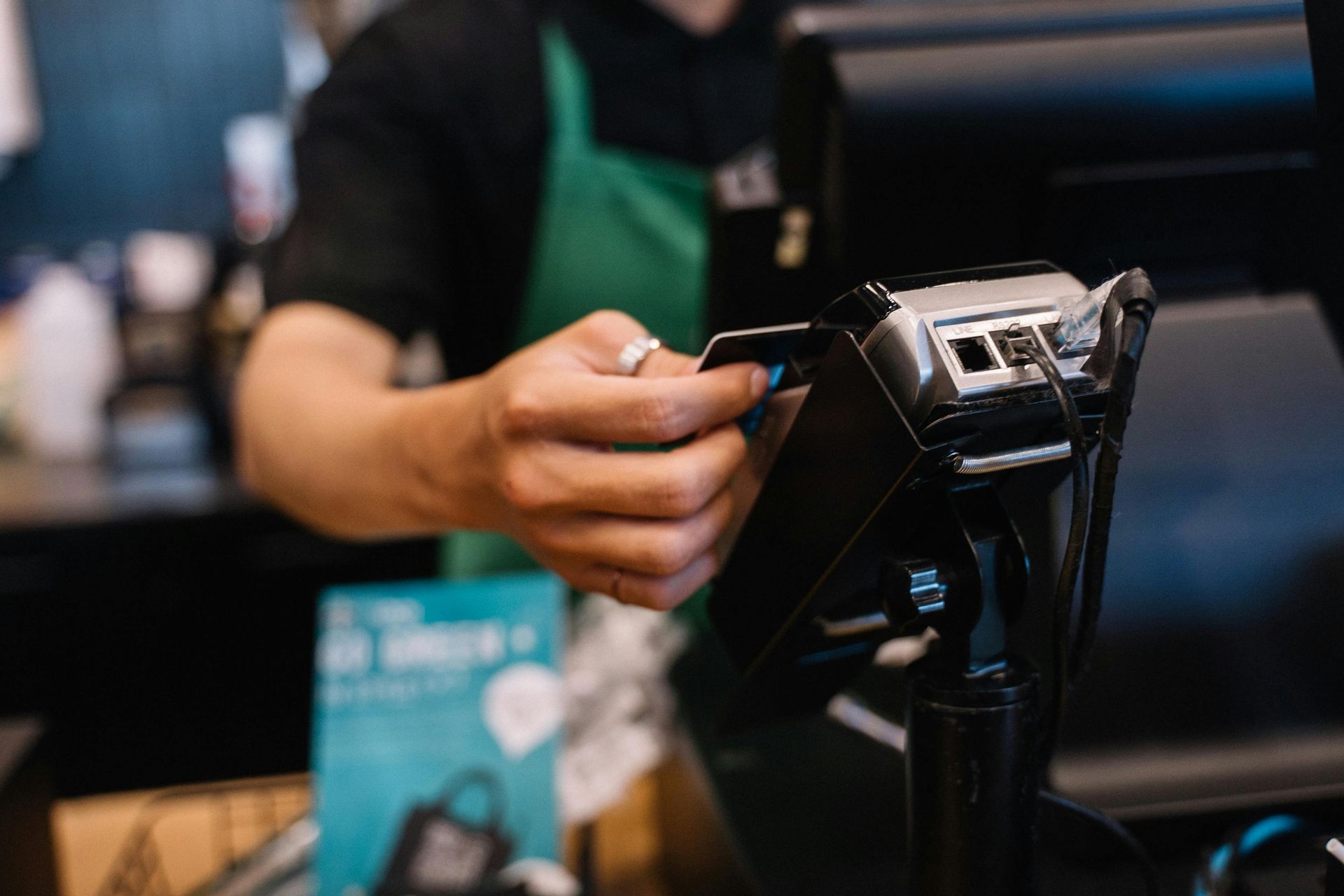 Person in green apron uses a credit card reader in a coffee shop setting.
