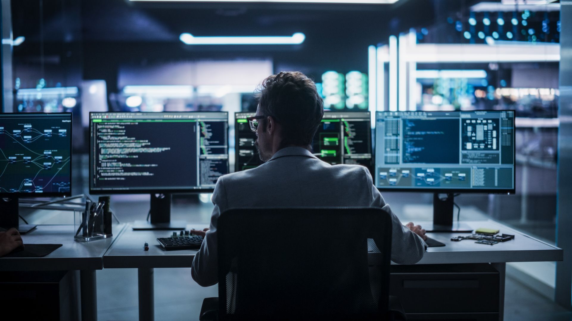 Man in a white coat working at a desk with three computer monitors displaying code and data in a dimly lit room.